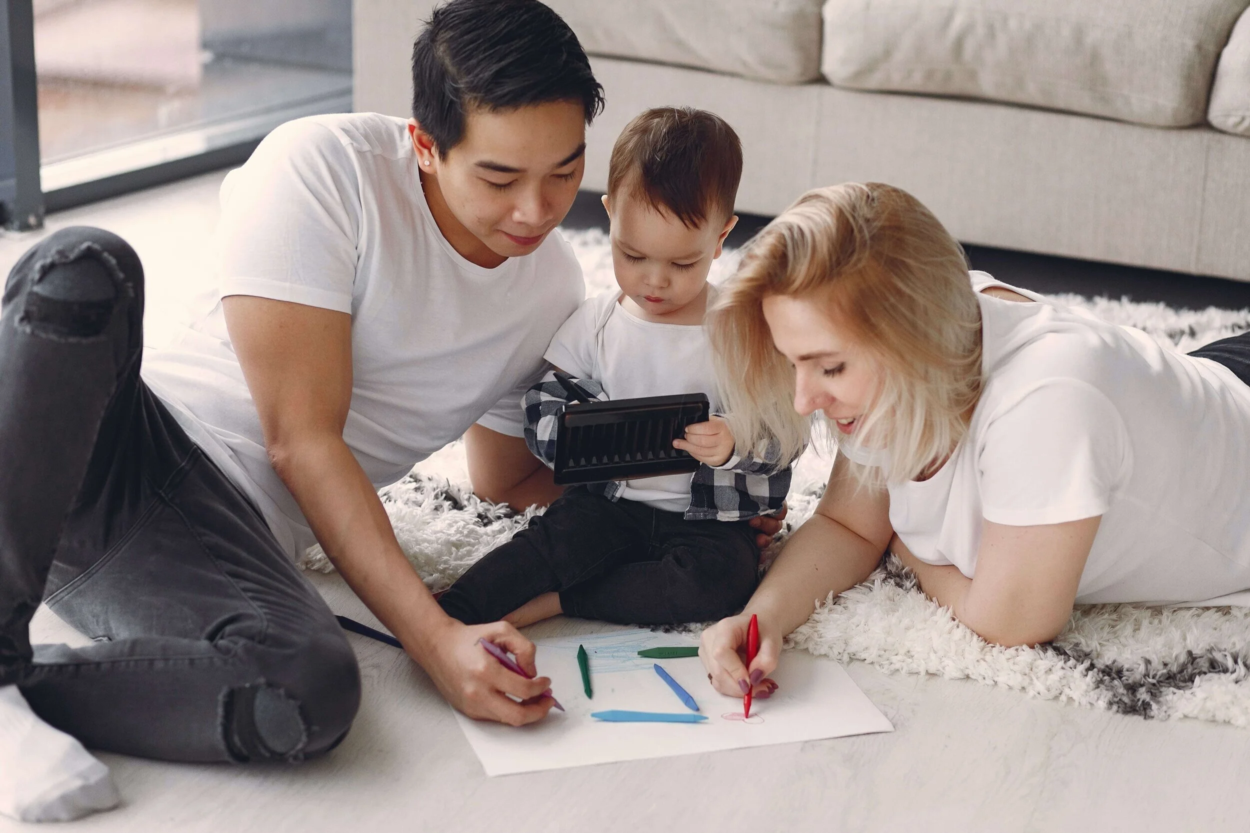 Three people, two adults and a child, are sitting on the floor with crayons and paper, drawing together in a bright living room.